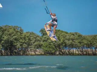 Osoba unosi się na kitecie nad wodą, tło stanowią drzewa i niebo. Logo KiteAway sugeruje niezapomniany wyjazd kitesurfingowy.