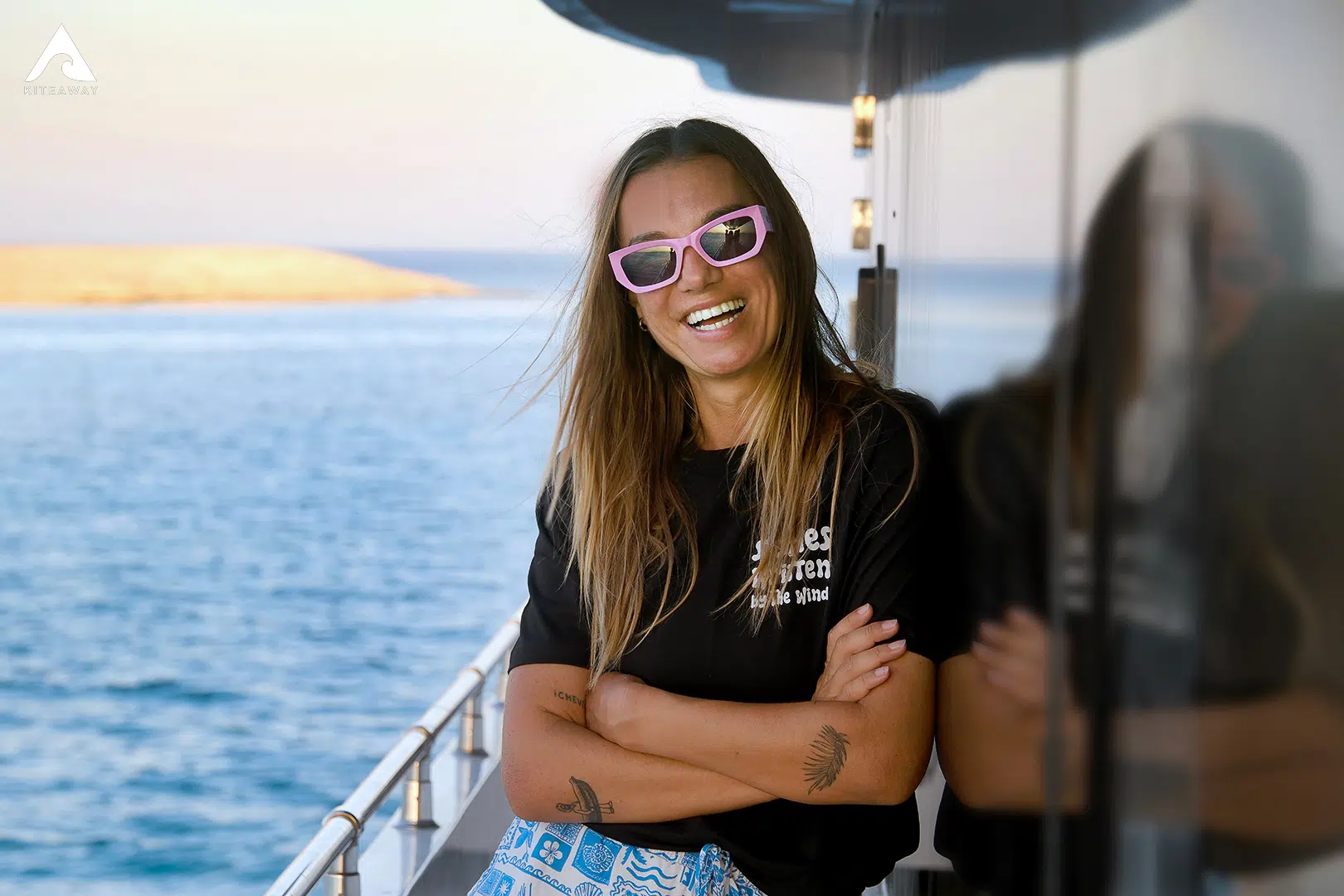 Kobieta z długimi włosami w okularach przeciwsłonecznych i swobodnych ubraniach stoi uśmiechnięta na pokładzie łodzi, gotowa do kitesurfingu w Brazylii. Ocean za nią.