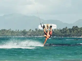Kitesurfer robi efektowny trik nad turkusową wodą na tle palm i gór - wakacje z kitesurfingiem z KiteAway.