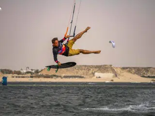 Rider wykonuje trik nad wodą podczas wyjazdu kitesurfingowego KiteAway; w tle plaża i inni uczestnicy kite campu.