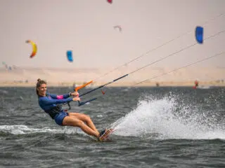 Uśmiechnięta kobieta w niebieskiej piance pływa na kite campie KiteAway; kolorowe latawce, kurs kitesurfingu w tle.