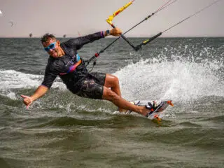 Kitesurfer w okularach i piance na KiteAway kite trip, efektowny splash; w tle wakacje z kitesurfingiem, kilku riderów.