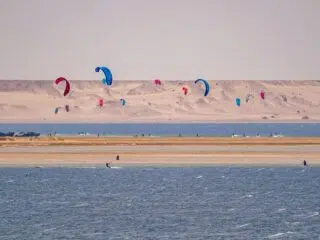 Kitesurferzy ślizgają się po wodzie z kolorowymi latawcami; w tle wydmy - idealny spot na wyjazdy kitesurfingowe z KiteAway.