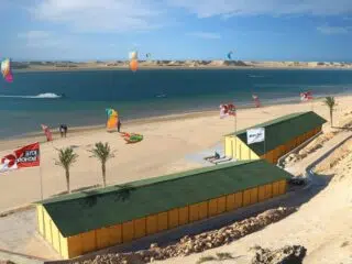 Słoneczna plaża KiteAway przy palmach i żółtych budynkach; kurs kitesurfingu na turkusowej wodzie - idealny kite camp.