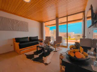 Przytulny salon z drewnianym sufitem, dużymi oknami i miejscem do relaksu po kite campie KiteAway; idealny na wyjazdy kitesurfingowe.