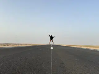 Osoba w pozie X na pustej drodze, pod błękitnym niebem, symbolizuje wolność na wyjeździe kitesurfingowym KiteAway.