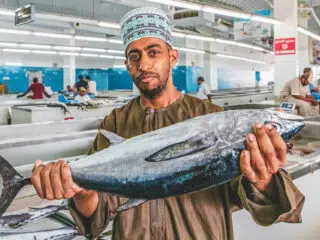 Mężczyzna w tradycyjnym stroju trzyma rybę na targu, obok ekipa KiteAway wspomina wyjazdy kitesurfingowe w Egipcie.