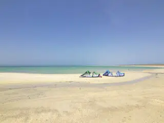 Szeroka, piaszczysta plaża z trzema kolorowymi latawcami kite nad turkusową wodą - idealny spot na wyjazdy kitesurfingowe z KiteAway.