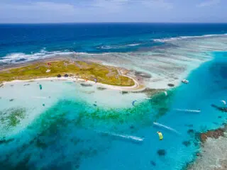 Mała zielona wyspa w turkusowej wodzie; wokół latający kitesurferzy, łódki i idealny spot na kurs kitesurfingu z KiteAway.