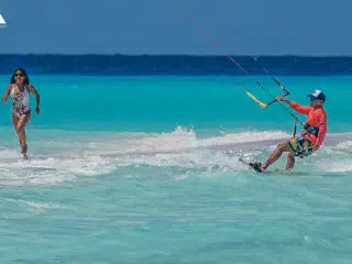Mężczyzna na kitesurfingu w turkusowej wodzie, kobieta biegnie plażą - wakacje z kitesurfingiem KiteAway, kite camp.