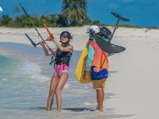 Uśmiechnięta kobieta w kasku i kamizelce na kursie kitesurfingu KiteAway; kite trip na plaży pod palmami, mężczyzna z foil boardem.