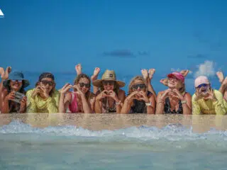 Siedem kobiet na brzegu wody, uśmiechnięte, w kolorowych strojach. Wakacje z kitesurfingiem z KiteAway - wyjazdy kite.