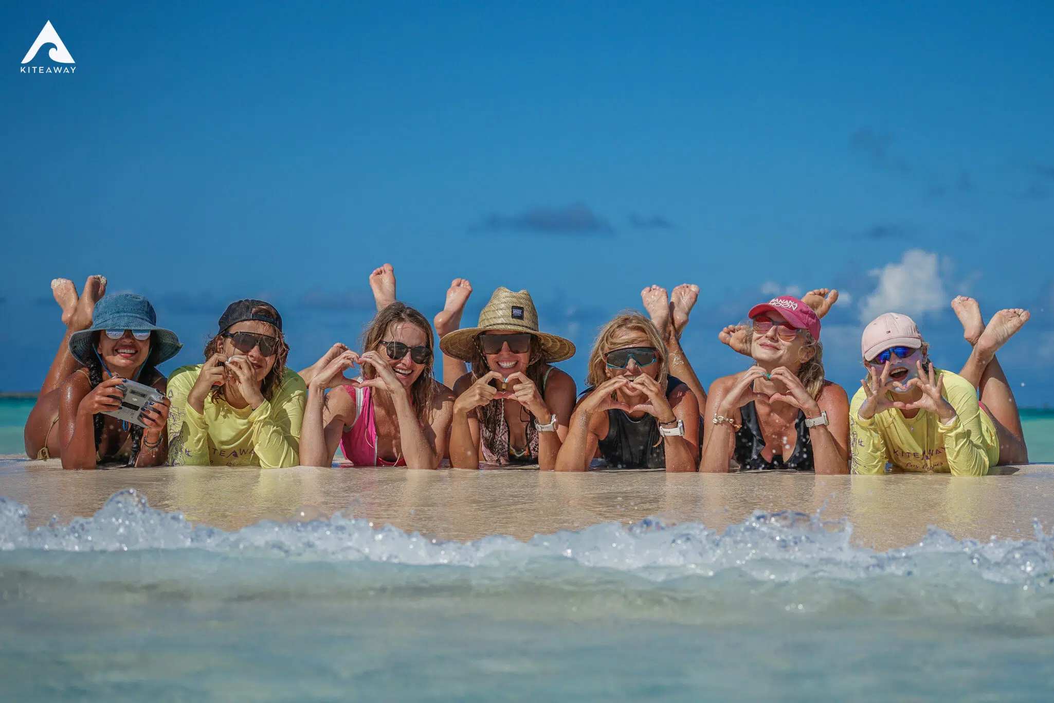 Siedem kobiet na brzegu wody, uśmiechnięte, w kolorowych strojach. Wakacje z kitesurfingiem z KiteAway - wyjazdy kite.