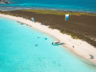 Plaża z białym piaskiem, turkusową wodą i kitesurferami z niebieskimi latawcami - idealne miejsce na wyjazdy kitesurfingowe z KiteAway.