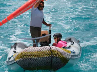 Mężczyzna na dmuchanej łodzi z żaglem, obok dziecka w kasku - idealne wakacje z kitesurfingiem z KiteAway na lazurowej wodzie.