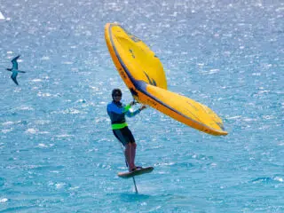 Rider na hydrofoilu z żółtym skrzydłem na turkusowej wodzie; idealny spot na wyjazdy kitesurfingowe z KiteAway.