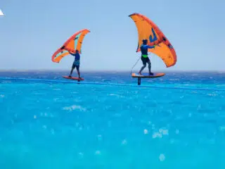 Dwie osoby na deskach hydrofoil z pomarańczowymi skrzydłami na lazurowej wodzie; idealne miejsce na wyjazdy kitesurfingowe KiteAway.
