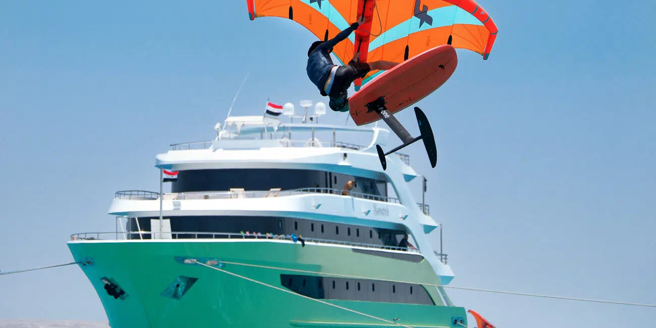 Rider na desce wodolot z pomarańczowym skrzydłem nad turkusową wodą i jachtem - idealne wakacje z kitesurfingiem KiteAway.