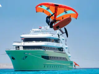 Rider na desce wodolot z pomarańczowym skrzydłem nad turkusową wodą i jachtem - idealne wakacje z kitesurfingiem KiteAway.