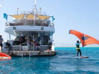 Osoba na foil boardzie z pomarańczowym skrzydłem przy łodzi; inni szykują sprzęt na wakacje z kitesurfingiem KiteAway.