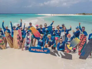 Uśmiechnięta grupa w niebieskich koszulkach z deskami na plaży promuje wyjazdy kitesurfingowe KiteAway na słonecznym kite spocie.