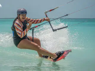 Kitesurfer w kasku i kolorowej bluzie pędzi po turkusowej wodzie Los Roques, uśmiecha się, trzyma latawiec, wzbijając fale.