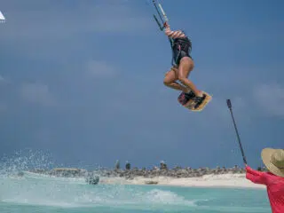 Kitesurfer w powietrzu nad turkusową wodą, filmowany podczas wyjazdu kitesurfingowego KiteAway; plaża i niebo w tle.