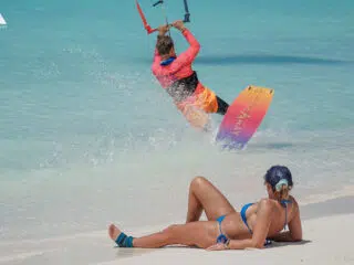 Kitesurfing w Los Roques: czysta turkusowa woda, słoneczne niebo, kobieta w niebieskim bikini obserwuje kitesurfera z piaszczystej plaży.
