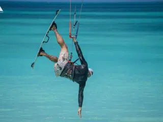 Kitesurfer w czapce i uprzęży wykonuje akrobację nad turkusową wodą Los Roques, dotykając ręką tafli i wysuwając nogi w powietrzu.
