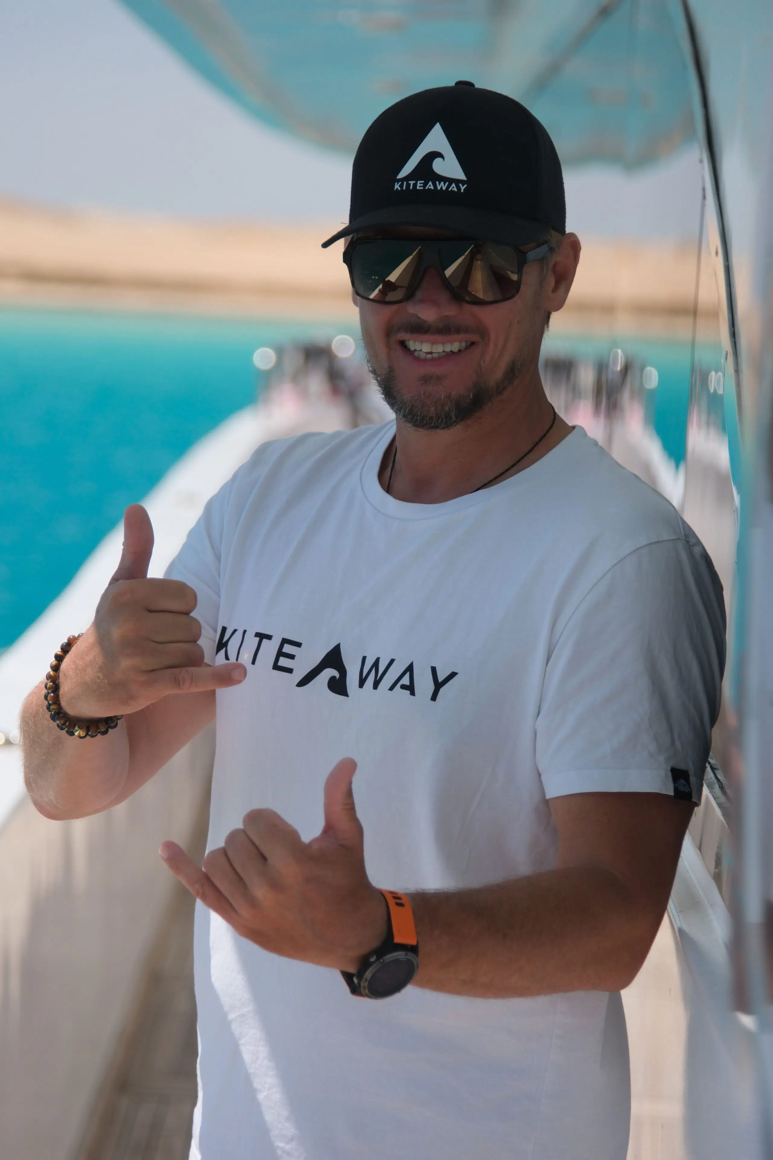 Uśmiechnięty mężczyzna w koszulce KiteAway pokazuje shaka na łodzi podczas wyjazdu kitesurfingowego nad turkusową wodę.