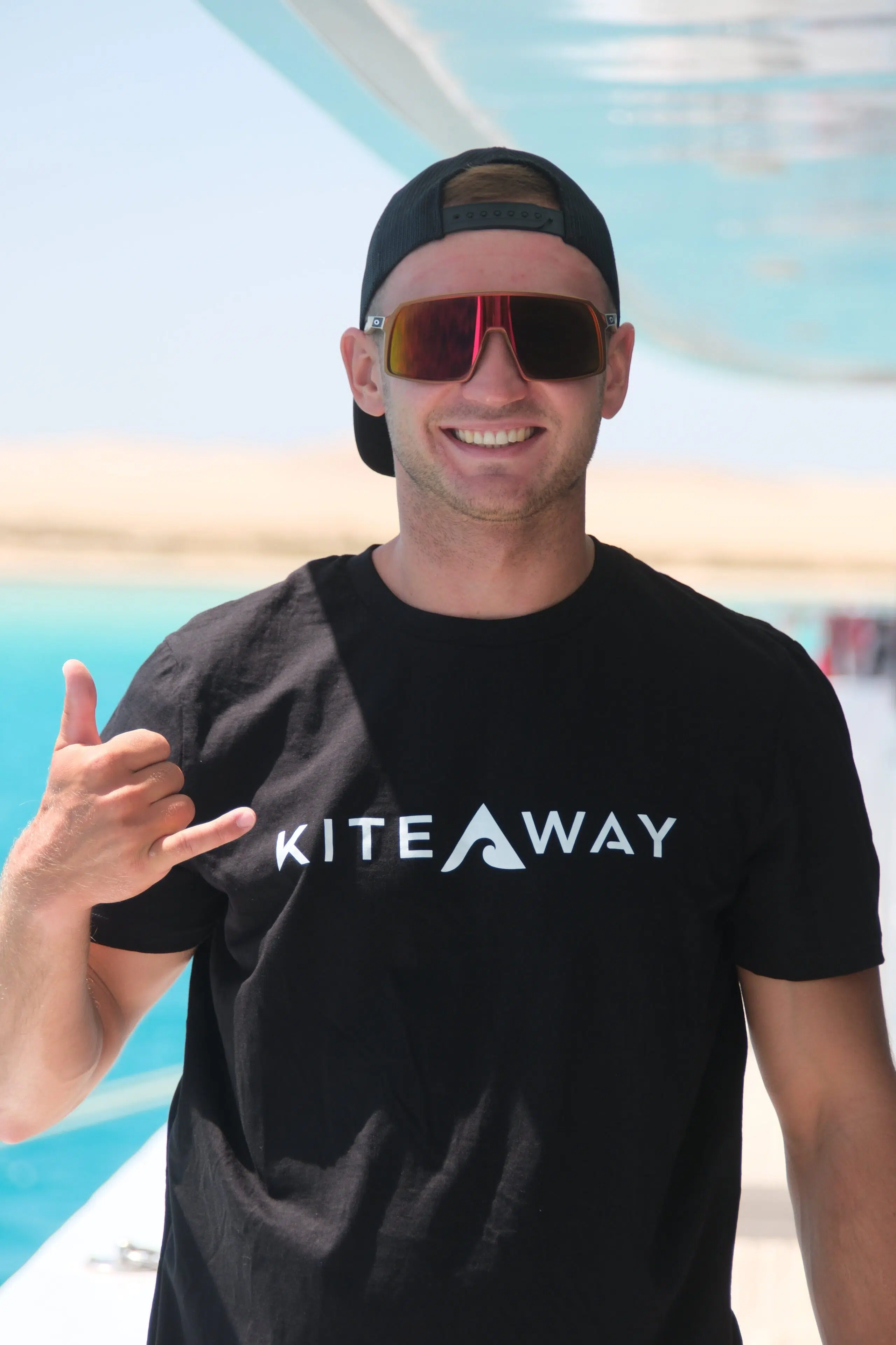 Uśmiechnięty mężczyzna w koszulce KiteAway na łodzi, shaka, turkusowa woda - gotowy na wyjazd kitesurfingowy z KiteAway.