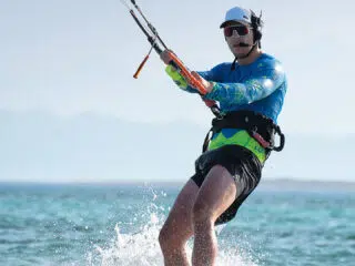 Uczestnik wyjazdu kitesurfingowego KiteAway pływa na desce, trzymając bar, na tle gór i nieba. Wakacje z kitesurfingiem.