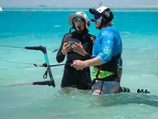 Dwie osoby w piankach podczas kursu kitesurfingu KiteAway stoją w turkusowej wodzie na kite campie. Słoneczna, tropikalna sceneria.