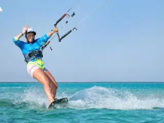 Uśmiechnięta osoba w niebieskiej koszulce kiteboarduje na turkusowej wodzie - wyjazdy kitesurfingowe z KiteAway.