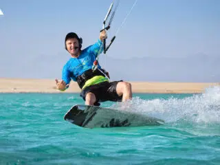 Uśmiechnięty kitesurfer na kite tripie KiteAway, pływa po turkusowej wodzie z widokiem na plażę i góry. Wakacje z kitesurfingiem.