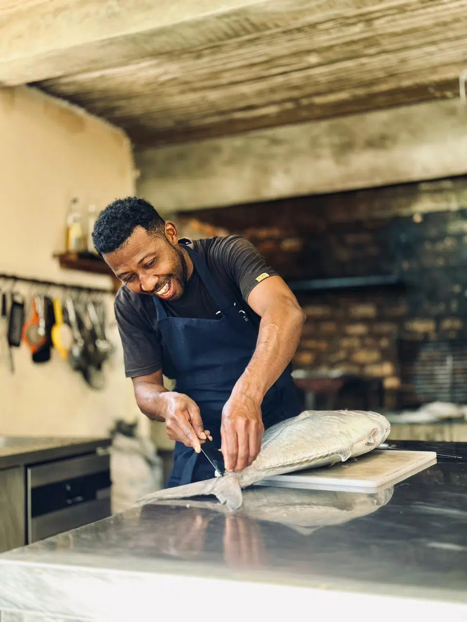 Uśmiechnięty mężczyzna w fartuchu filetującym rybę w kuchni po dniu pełnym wrażeń na kite camp z KiteAway - idealny posiłek po wyjazdach kitesurfingowych.