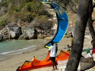 Plaża pełna zieleni i klifów, osoba z niebiesko-pomarańczowym latawcem szykuje się do kursu kitesurfingu z KiteAway.