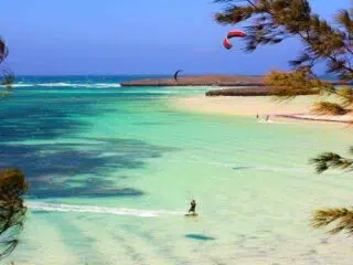 Kitesurfer pływający po czystej turkusowej wodzie w pobliżu piaszczystej plaży Madagaskaru, z drzewami z przodu, innym kitesurferem i błękitnym niebem.