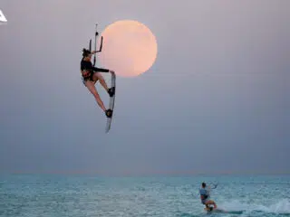 KiteAway: kobieta unosi się z latawcem na tle pełni księżyca - kurs kitesurfingu, wakacje z kitesurfingiem, drugi kitesurfer w tle.