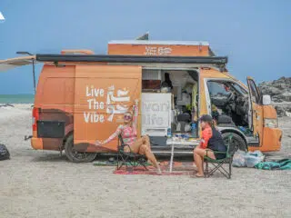 Dwie osoby przy stole pod markizą obok pomarańczowego kampera na plaży - idealny kite camp KiteAway, wakacje z kitesurfingiem.