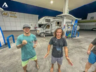 Dwóch uśmiechniętych mężczyzn na stacji benzynowej przy kamperze nocą, w trakcie wyjazdu kitesurfingowego KiteAway.