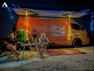 Dwie kobiety relaksują się przy pomarańczowym vanie na plaży po dniu na kite campie KiteAway; obok deska SUP i napoje.