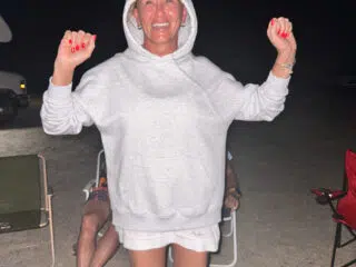 Uśmiechnięta kobieta w szarej bluzie na plaży podczas kite campu KiteAway - wakacje z kitesurfingiem, w tle uczestnicy.