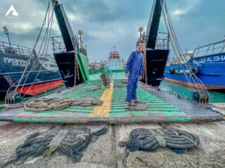 Mężczyzna w niebieskim kombinezonie na pokładzie statku w porcie, za nim przygotowania do kite trip z KiteAway pod zachmurzonym niebem.