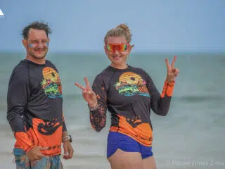 Dwie osoby w koszulkach KiteAway pozują na plaży przy oceanie, uśmiechnięte-idealny dzień na wyjazdy kitesurfingowe lub kite camp.