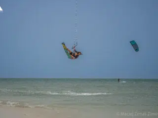 Kiteboarder skacze nad wodą blisko plaży podczas kursu kitesurfingu z KiteAway; w tle drugi uczestnik, słoneczny kite spot.