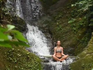 Kobieta w sportowym stroju medytuje na skale przy wodospadzie wśród zieleni, relaksując się przed wyjazdem kitesurfingowym z KiteAway.