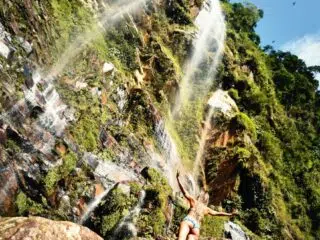 Osoba w stroju kąpielowym stoi na skałach u stóp zielonego klifu z wodospadem - idealne miejsce na wyjazdy kitesurfingowe z KiteAway.