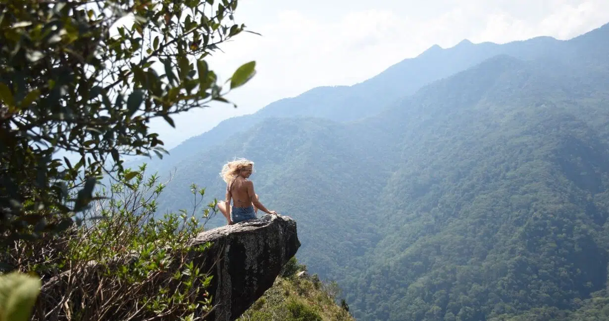 Osoba z długimi blond włosami siedzi na skale, patrząc na zielone góry - gotowa na kite trip z KiteAway, wakacje z kitesurfingiem.