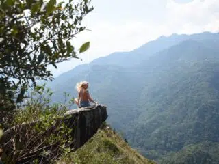 Osoba z długimi blond włosami siedzi na skale, patrząc na zielone góry - gotowa na kite trip z KiteAway, wakacje z kitesurfingiem.